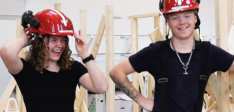 två elever med röda skyddshjälmar står och skrattar i bygghall